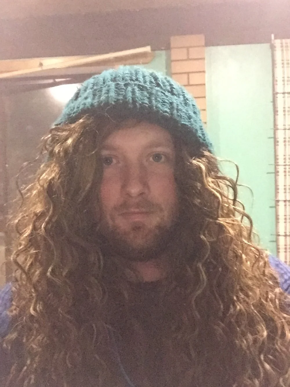 A man with long curly hair wearing a knitted beanie hat, inside a room with brick and wood walls.