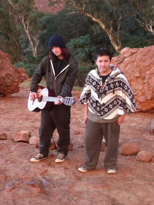 Two young boys standing outdoors on rocky terrain, one holding a guitar. One boy is wearing a black beanie, dark jacket, and jeans, and the other boy is wearing a patterned poncho and gray pants. Trees and large rocks are in the background.