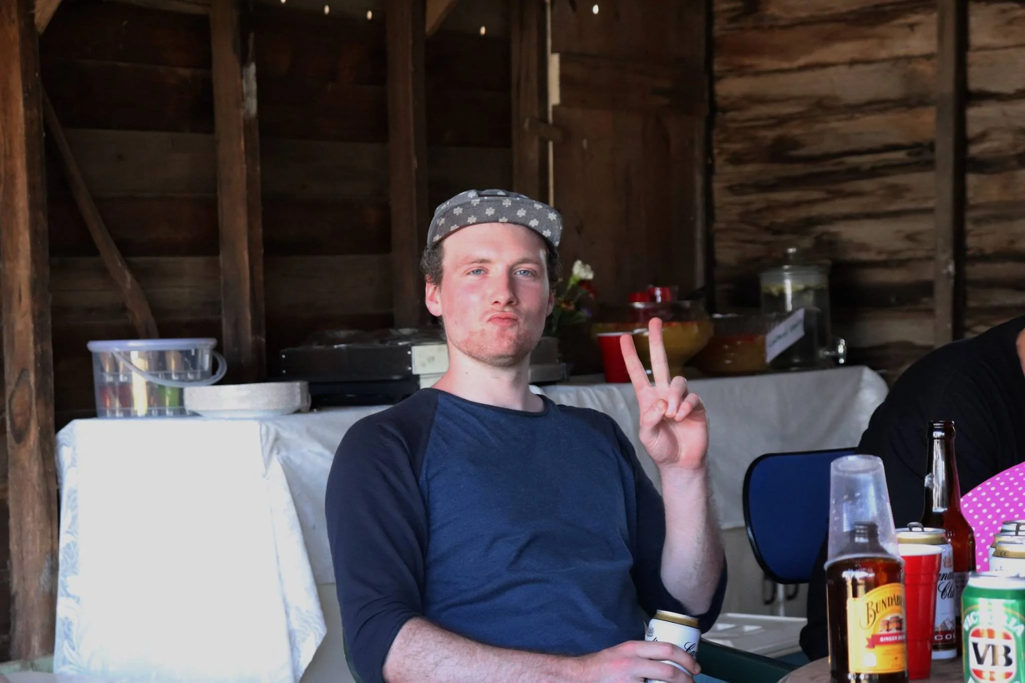 A young man with light skin, wearing a gray patterned cap and a dark blue long-sleeve shirt, making a peace sign with his right hand while holding a drink can in his left hand. He is seated at a table with various beverages, in a rustic wooden settin