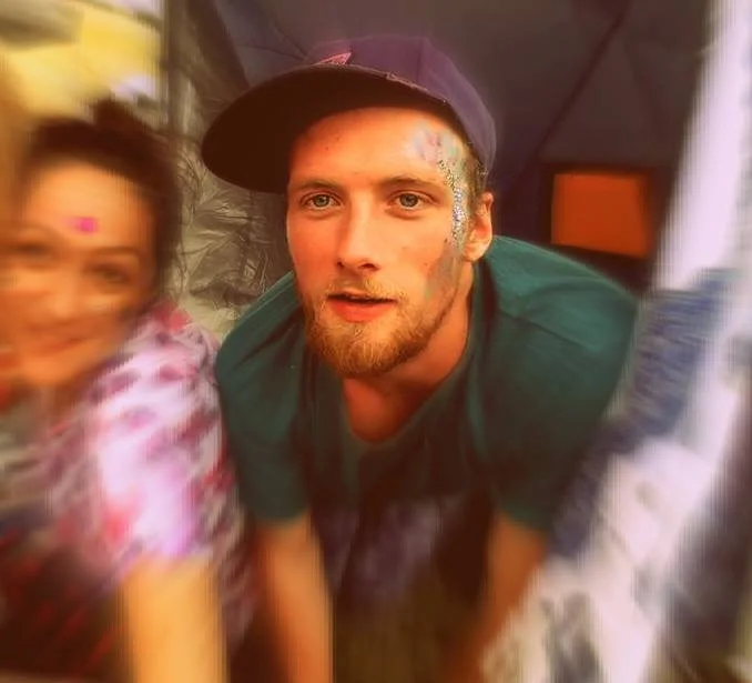 A young man with colorful paint on his face and a beard, wearing a baseball cap, looking at the camera. Blurred woman next to him, in a colorful outfit, also with paint on her face, smiling and slightly out of focus.