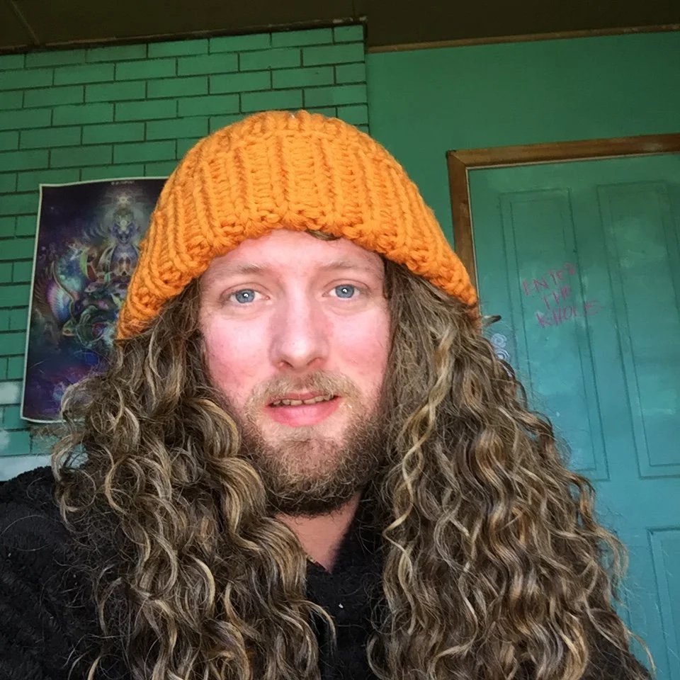 A man with long curly hair, a beard, wearing an orange knitted hat, inside a room with green brick walls and a teal door in the background.