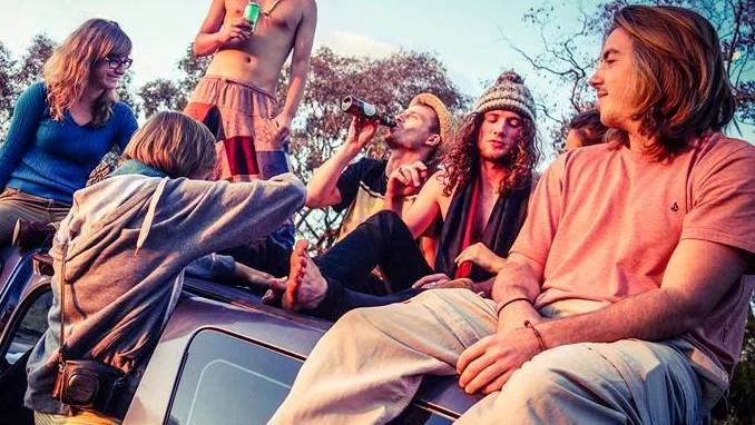 A group of young friends hanging out outdoors, sitting on and around a vehicle, with some drinking and chatting on a sunny day.