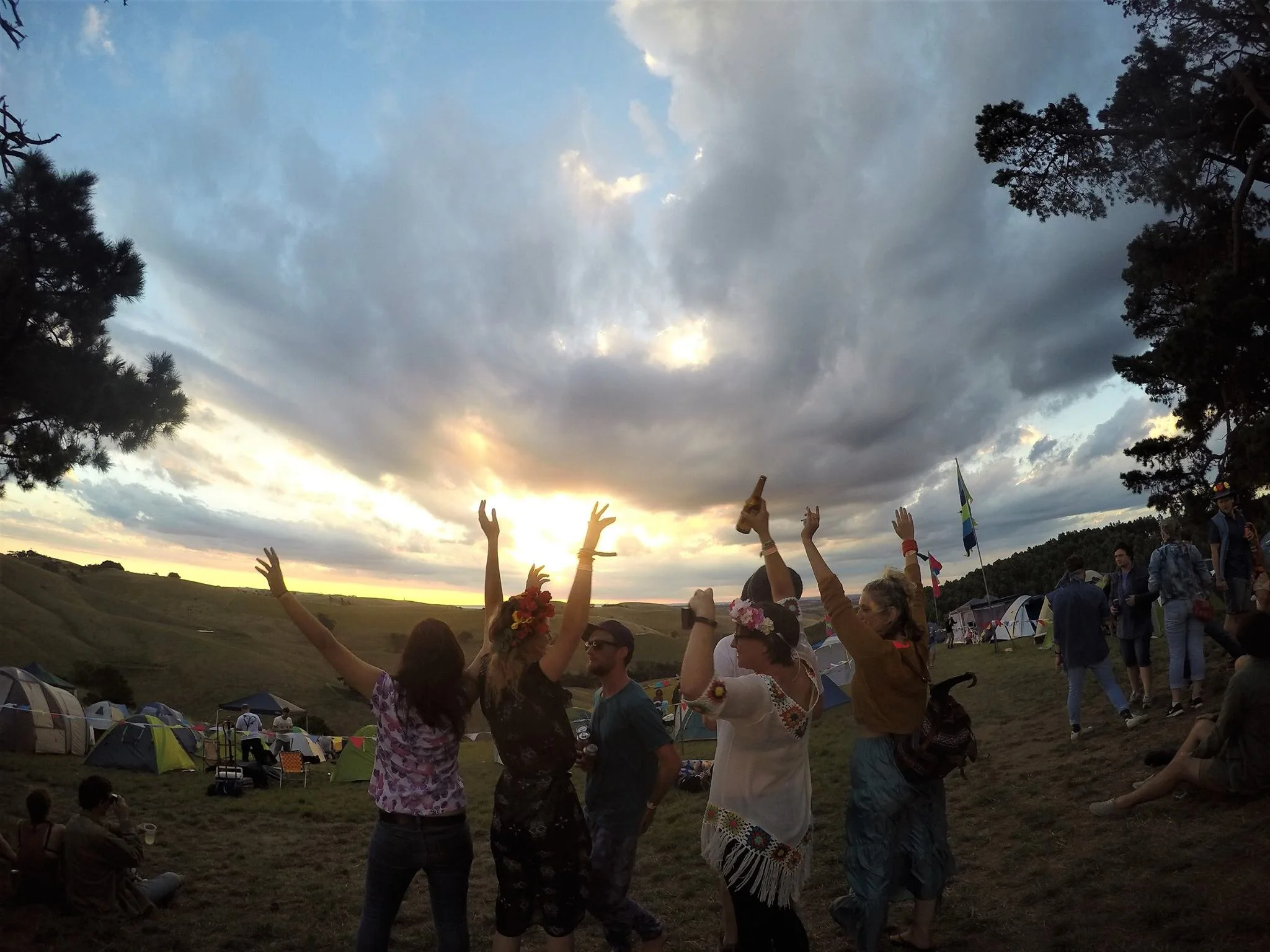 A group of people at an outdoor festival during sunset, dancing with arms raised, some wearing flower crowns, with tents and trees in the background under a partly cloudy sky.