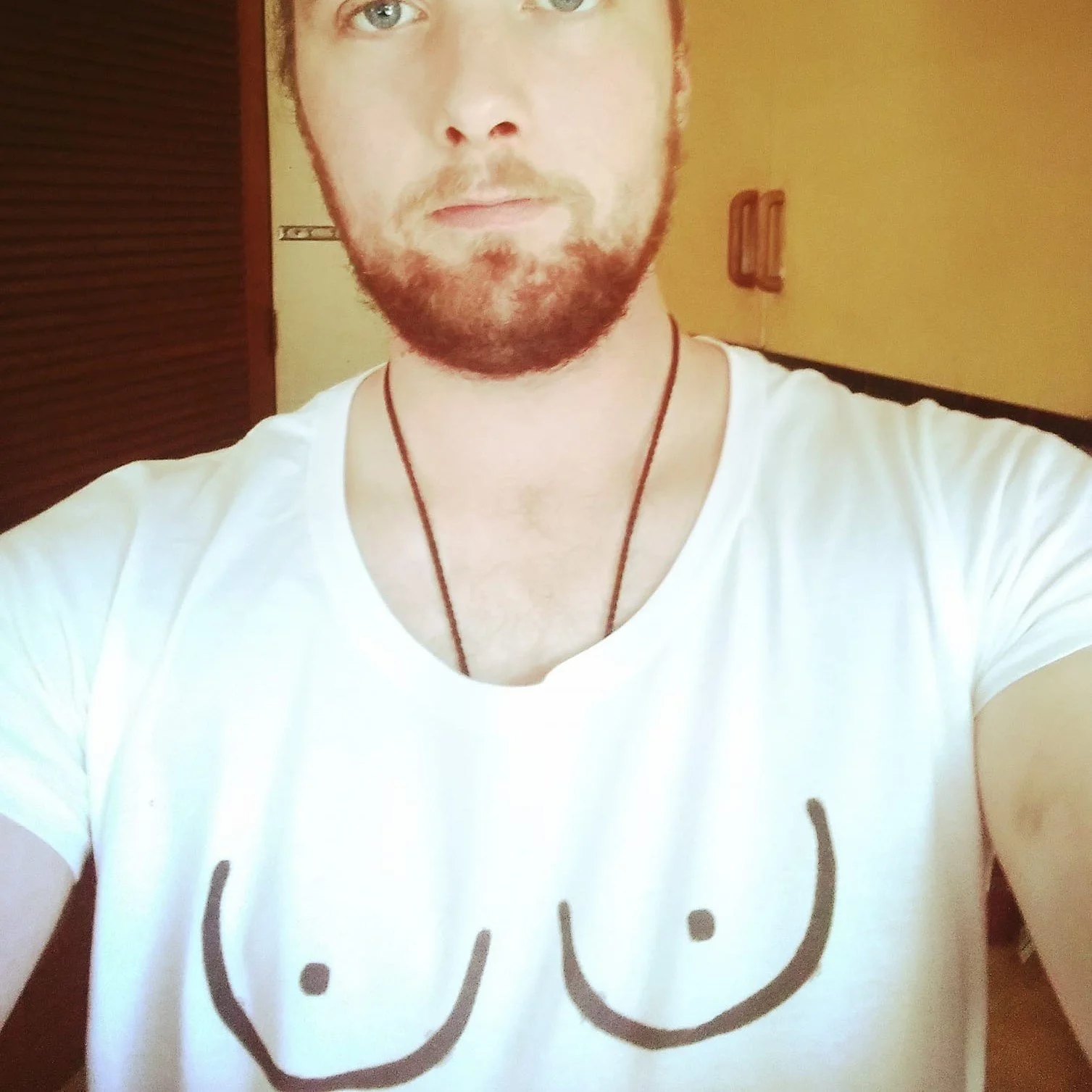 A person with a beard wearing a white T-shirt with a cheeky face drawing on it, and a black necklace, taking a selfie indoors.