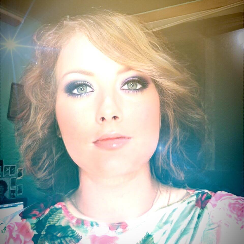 A woman with wavy red hair and makeup, wearing a floral top, looking slightly upward in an indoor setting with a light source creating a lens flare.