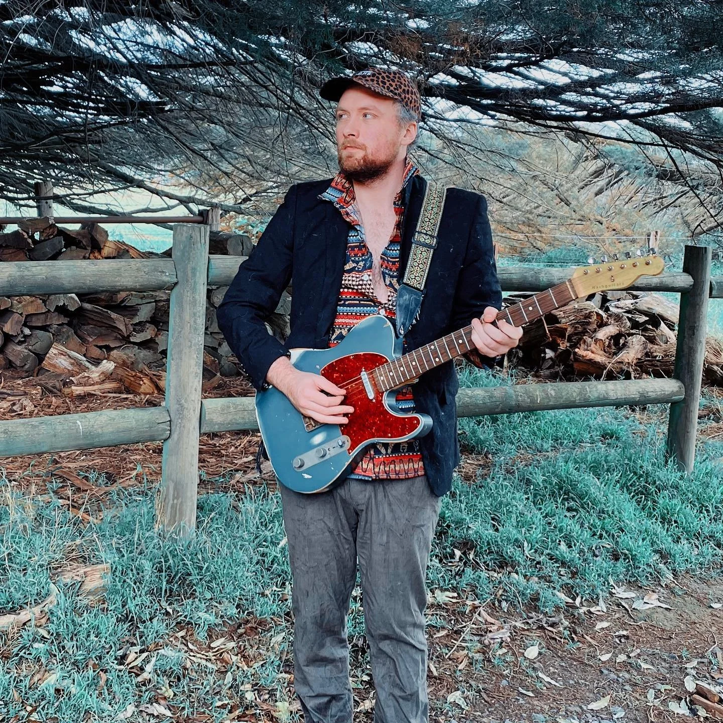 A man with a beard and a cap playing an electric guitar outdoors near a wooden fence and a pile of firewood, under a canopy of tree branches.