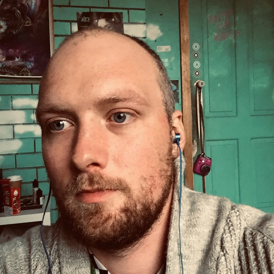 A close-up of a man with a beard and short hair, wearing blue earphones, in a room with a teal brick wall, a teal door with pegs and hanging objects, and posters on the wall.