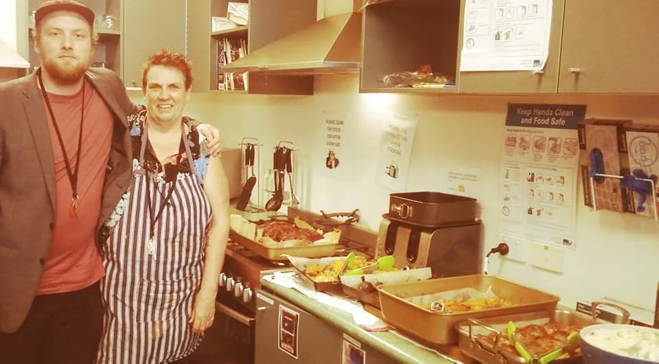 A man and woman standing in a kitchen with food trays and cooking appliances, smiling at the camera.