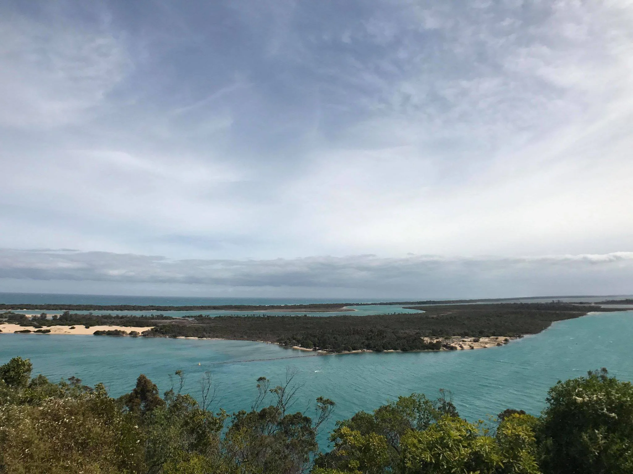 A landscape view of a large body of turquoise water with sandy and forested areas along the shoreline, under a partly cloudy sky.