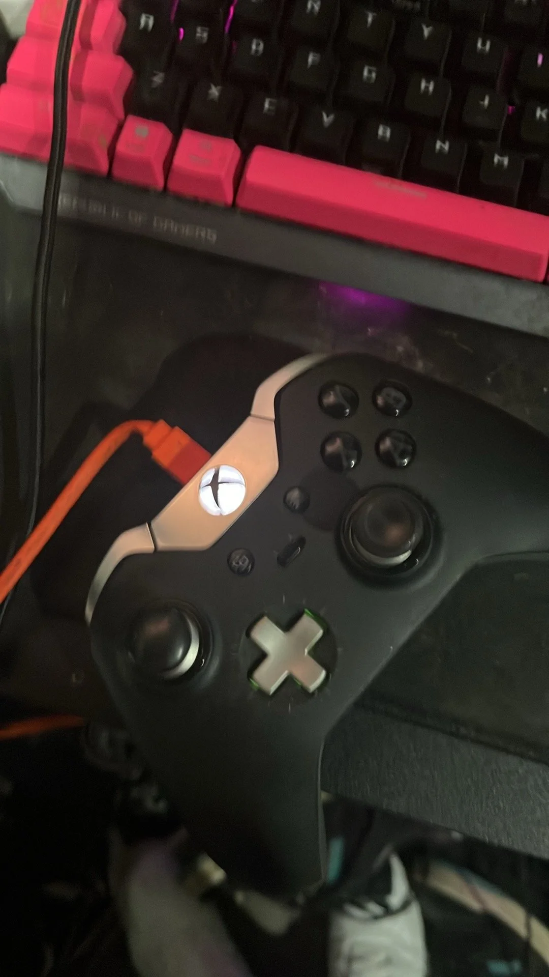 A gaming setup featuring a black Xbox controller, a mechanical keyboard with pink and black keys, and an orange gaming headset cable.
