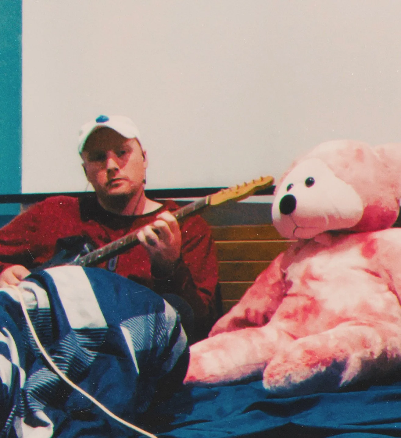 Person sitting on a bench playing guitar next to a large pink stuffed bear with a white face.