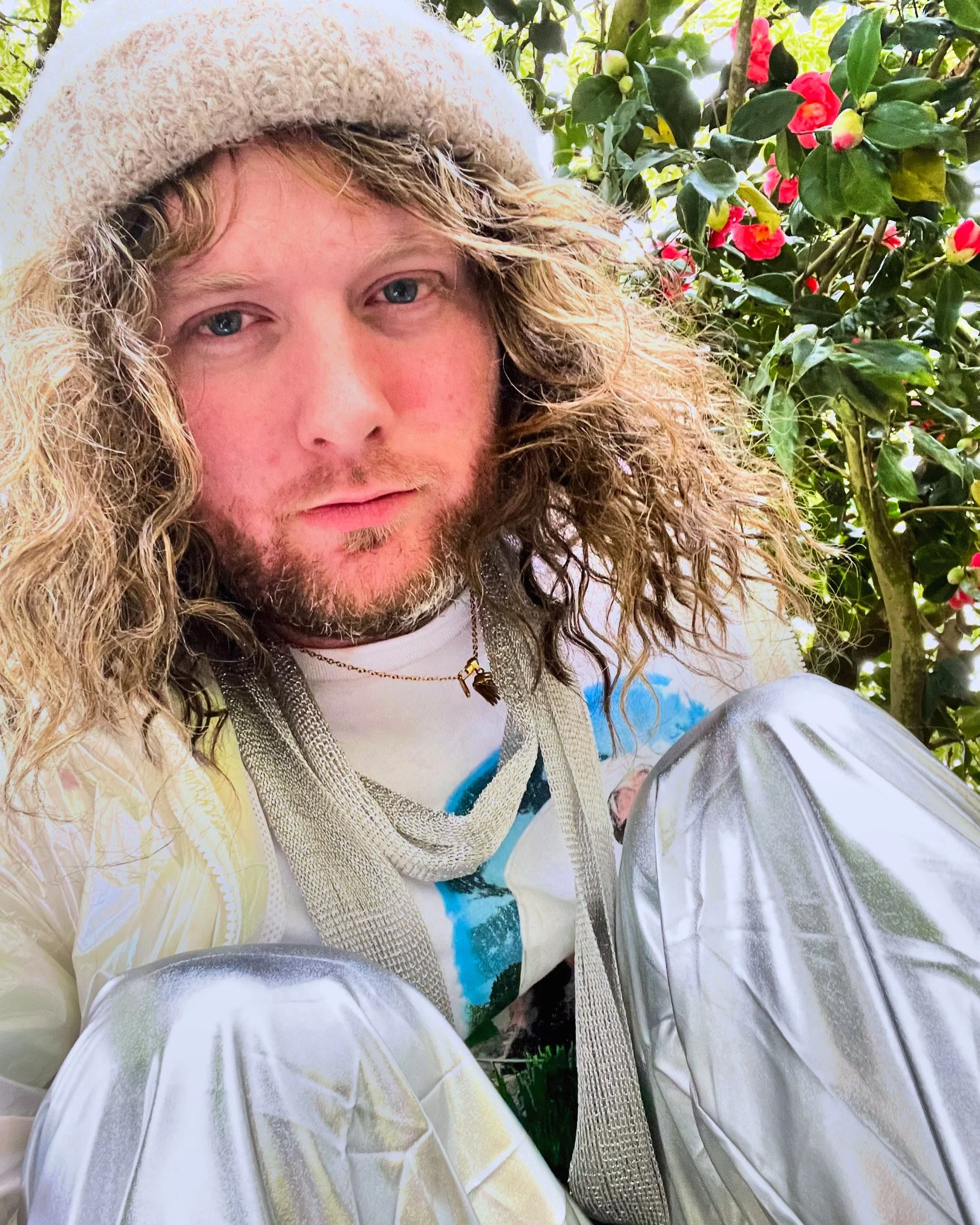 A young man with long curly hair and a beard, sitting outdoors near green foliage and red flowers, wearing a beige knit hat, a patterned shirt, and metallic-colored pants.
