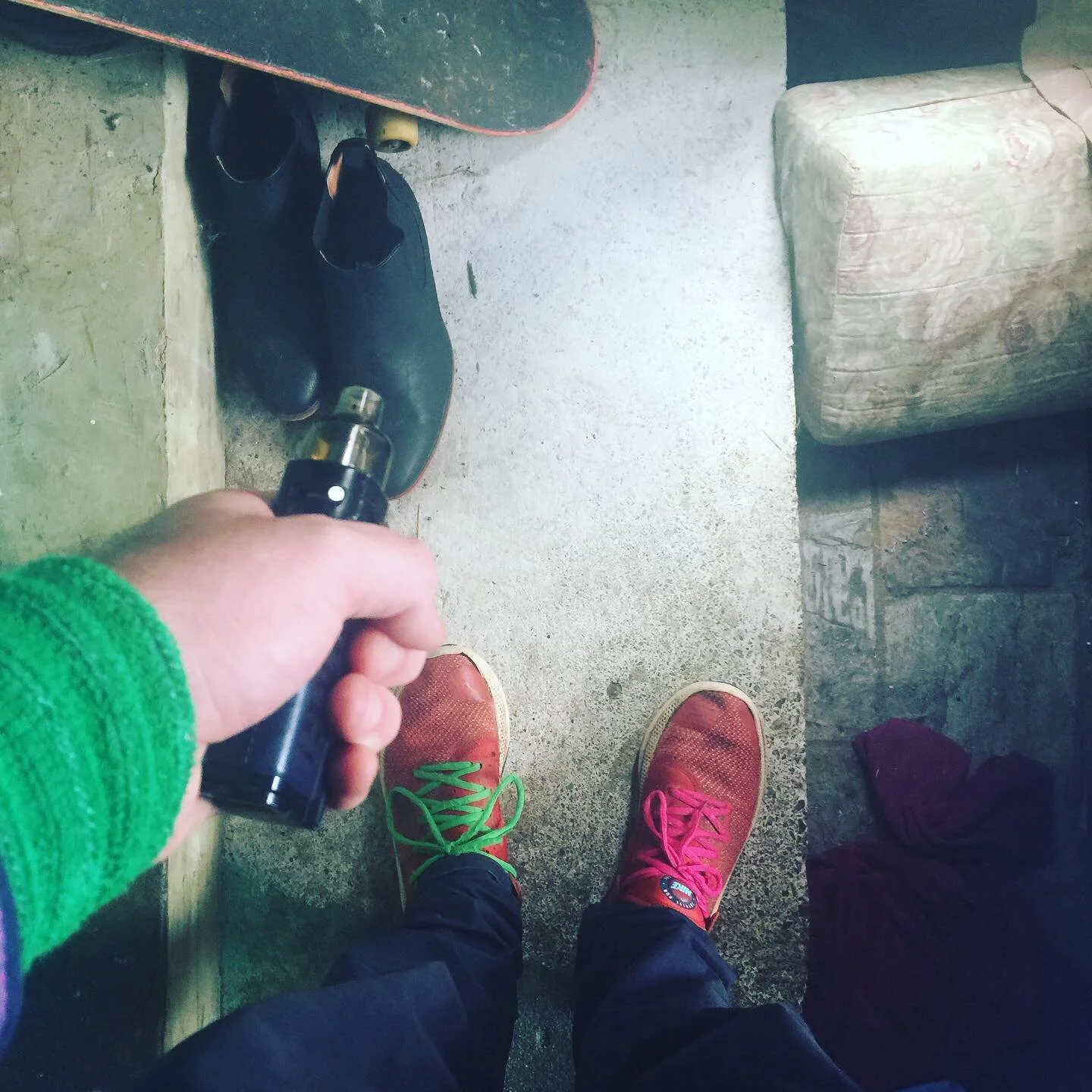 Person taking a photo of their colorful sneakers while standing on a concrete floor, with skateboard and shoes nearby, in an indoor setting.