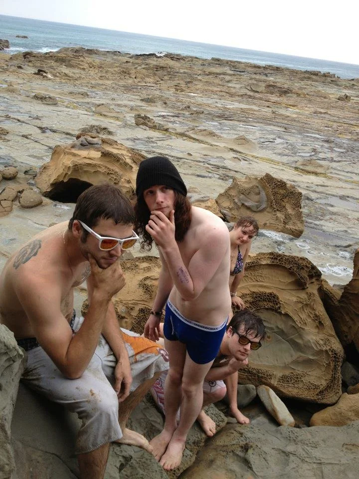 Four people at a rocky beach, two males in the foreground sitting on rocks, one wearing sunglasses and the other wearing a beanie and underwear. Two females in the background among rocks, one wearing glasses and the other with curly hair. The ocean i