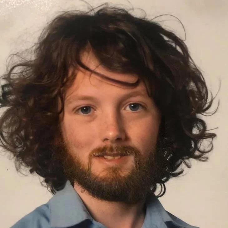 Portrait of a man with curly, shoulder-length hair, a beard, blue eyes, wearing a collared shirt.