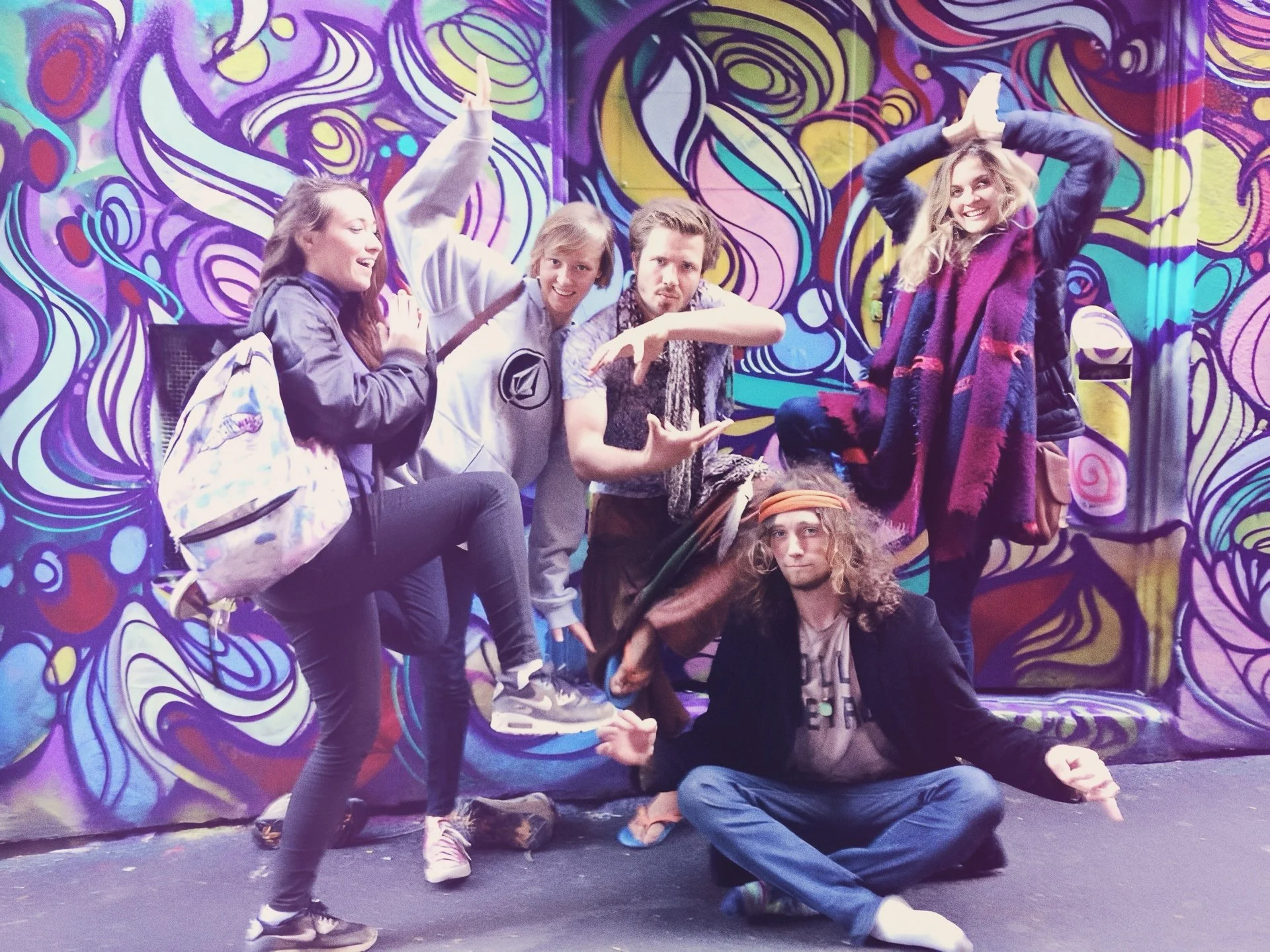 Group of six people posing playfully in front of a colorful, abstract mural on the wall.