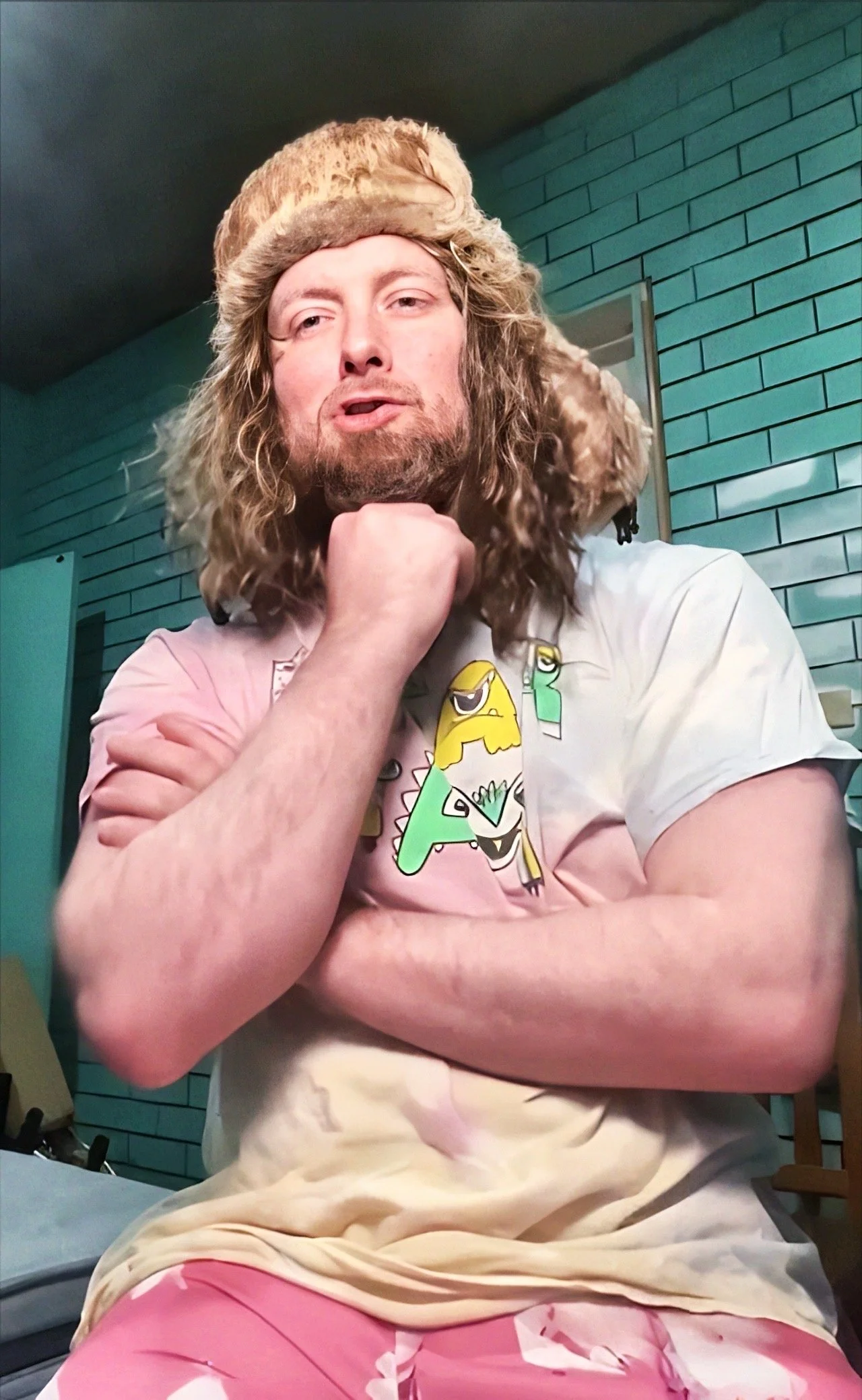 A man with long, curly blond hair and a beard wearing a fuzzy hat sits in front of a teal brick wall, resting his chin on his hand, wearing a colorful cartoon-themed t-shirt and pink pants.