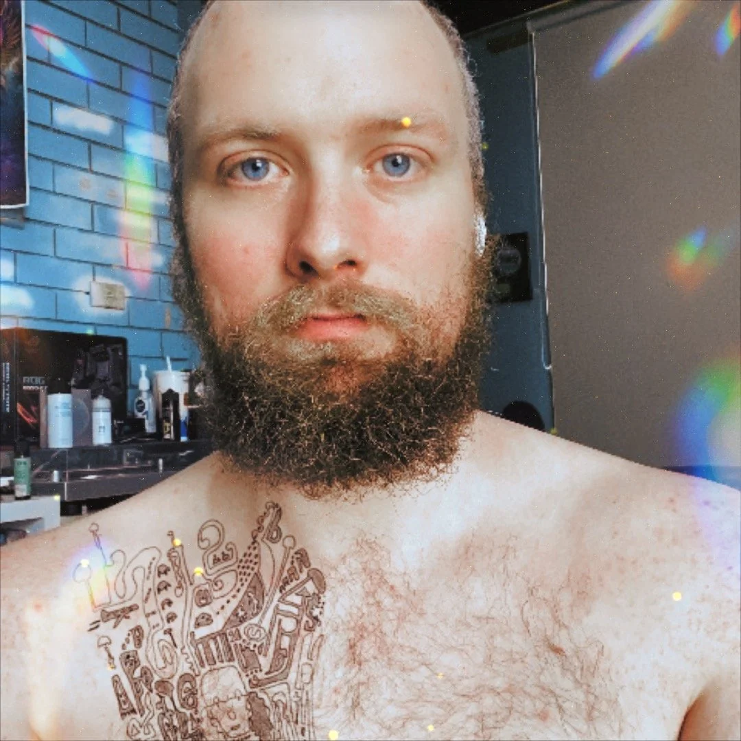 A shirtless man with a full beard and blue eyes takes a selfie in a room with a colorful brick wall. There are lots of grooming products on a counter behind him, and the room is lit with natural or colored light, creating rainbow-like spots.