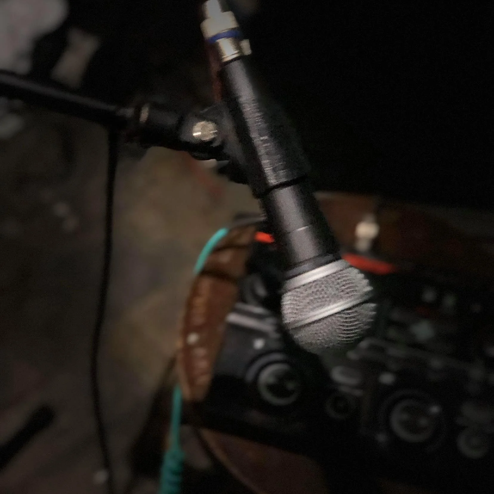 Close-up of a microphone mounted on a microphone stand, connected to audio equipment, with some wires and electronic devices in the background.