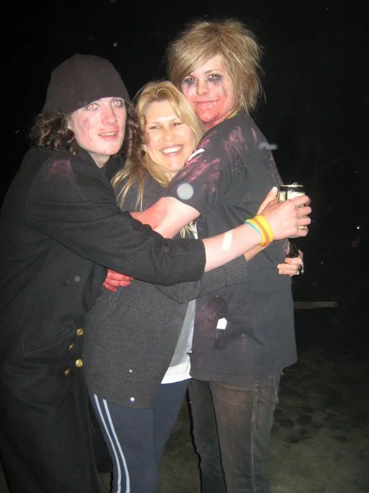 Three young people hugging, smiling, and celebrating at night. One person wears a black hoodie and beanie, another wears a gray sweater, and the third has messy blonde hair and is holding a drink.
