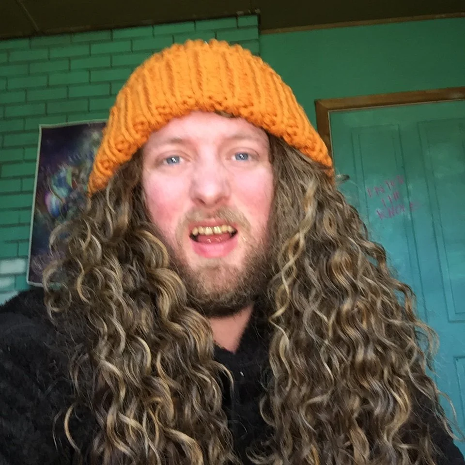 A person with long, curly hair wearing an orange knit beanie and a black top, smiling with their mouth slightly open.