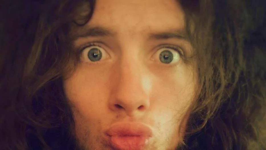 Close-up of a person with long, wavy hair, wide eyes, and pursed lips, looking directly at the camera.