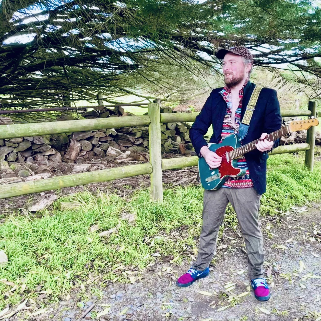 A man holding a guitar standing outdoors on a dirt path near a wooden fence and a pile of firewood, with green grass and trees overhead.