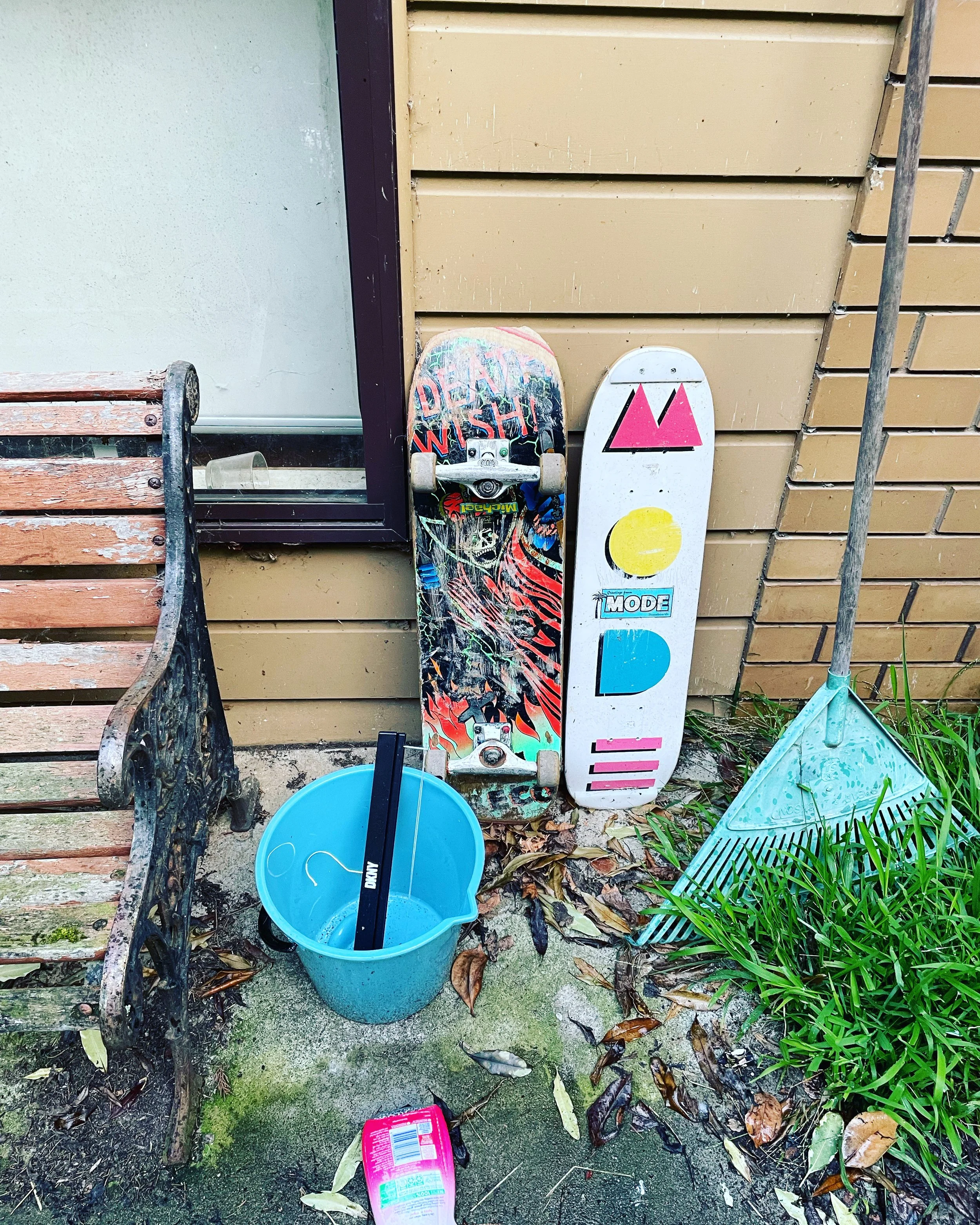 Two skateboards leaning against a tan wooden wall with a window, next to a rusty bench, a green rake, a blue bucket with a black item inside, and fallen leaves on the ground.