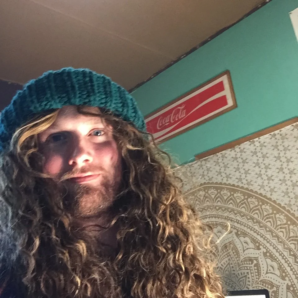 A person with long curly hair, wearing a knitted hat, taking a selfie indoors with a vintage Coca-Cola sign on the wall.