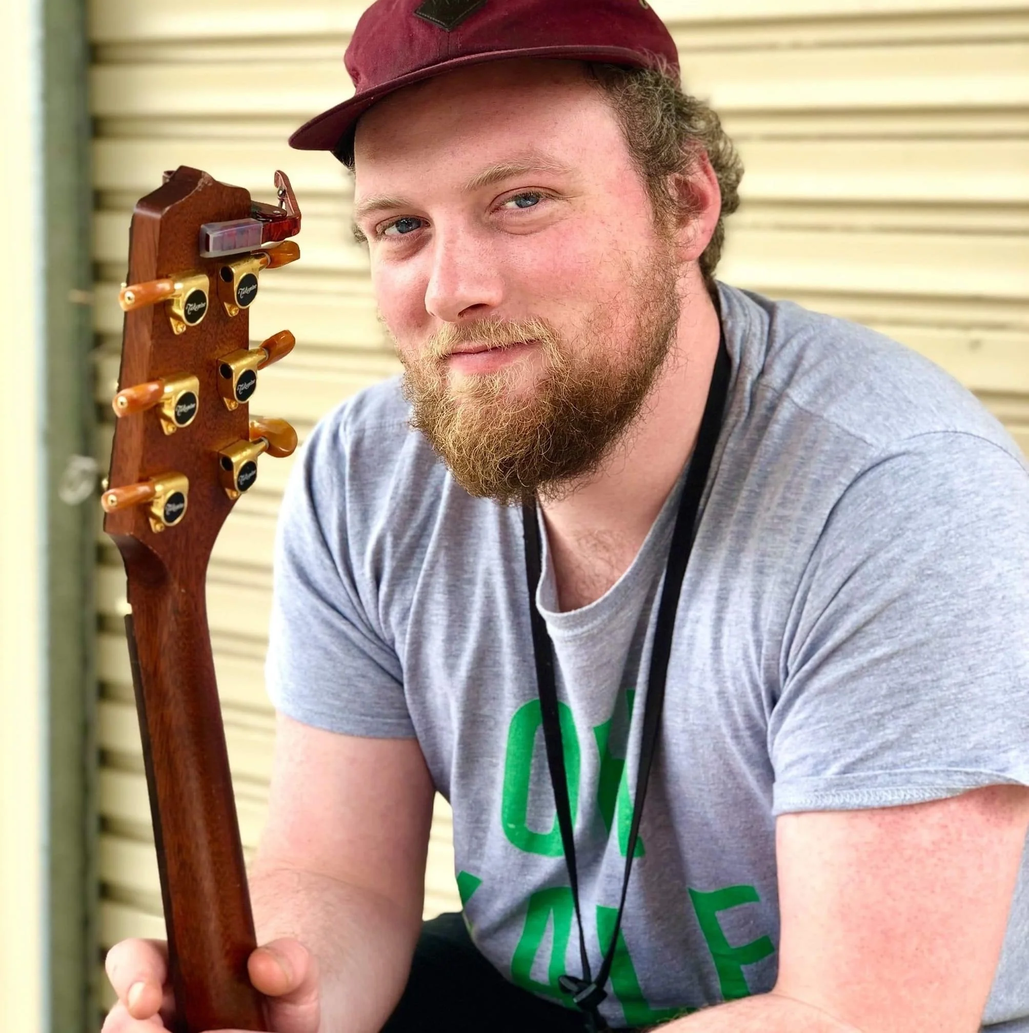 A man with a beard, wearing a maroon cap and a gray t-shirt, holding the neck of an acoustic guitar.