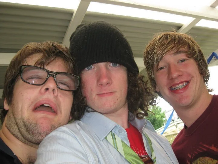Three young men taking a selfie outdoors under a roof with a cloudy sky and trees in the background.