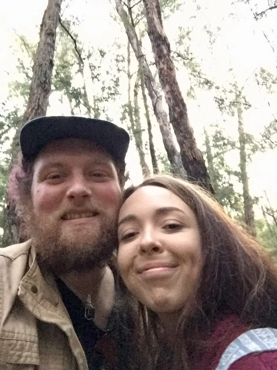Two people smiling in a forest with tall trees and sunlight filtering through the leaves.