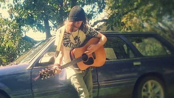 Person with long hair wearing a beanie and tie-dye shirt playing acoustic guitar next to a blue car in an outdoor setting with trees and sunlight.