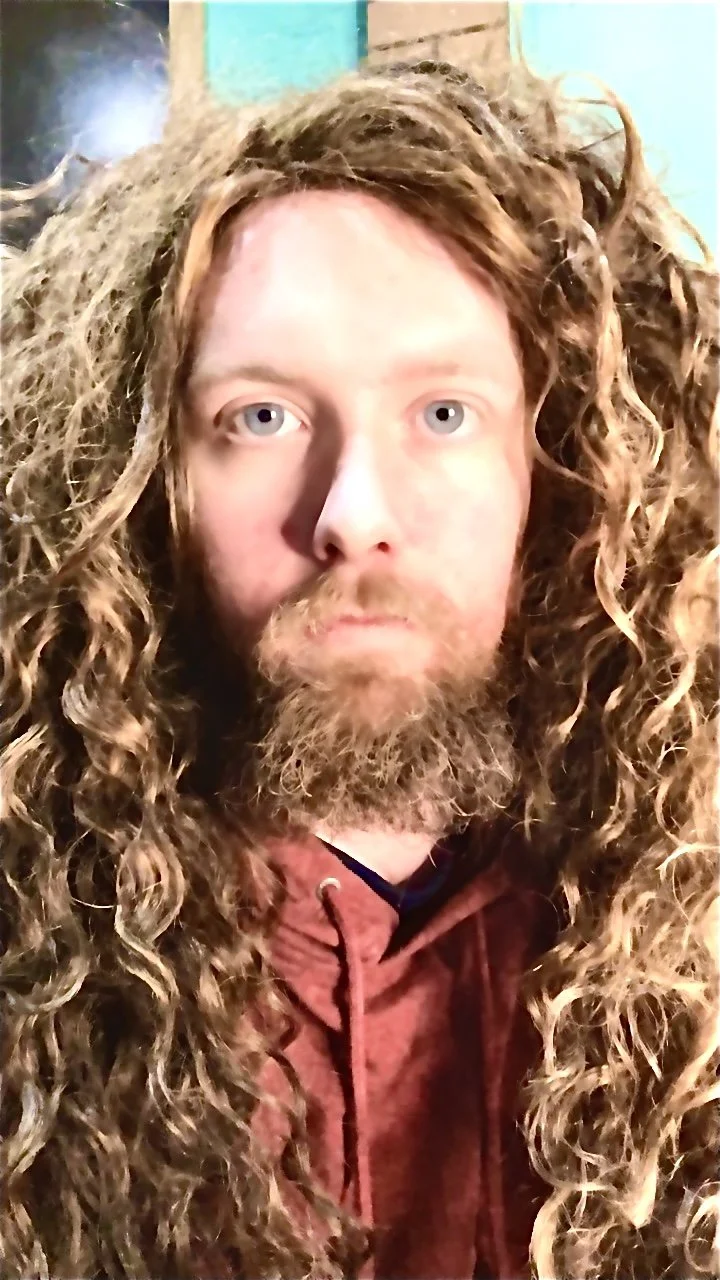 Close-up of a man with long, curly, light brown hair and a full beard wearing a red hoodie, looking directly at the camera indoors.