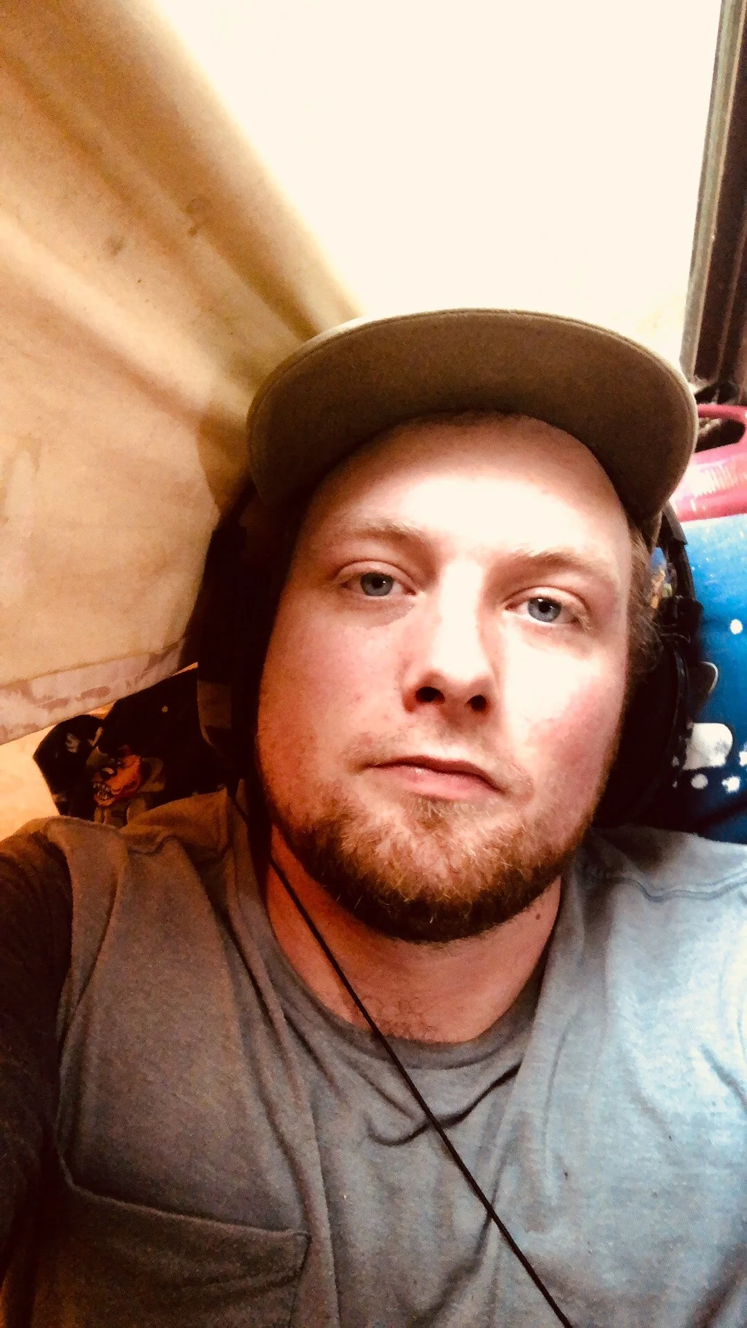 A man lying down on a bed, looking at the camera, wearing a gray shirt, a baseball cap, headphones, and a necklace.