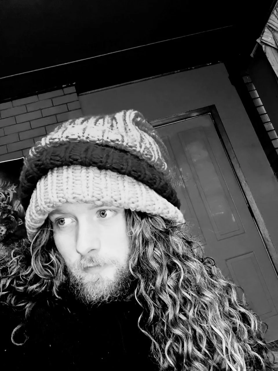 A young man with long curly hair and a beard, wearing a knitted beanie, looking to the side. Black and white photo, indoor setting.