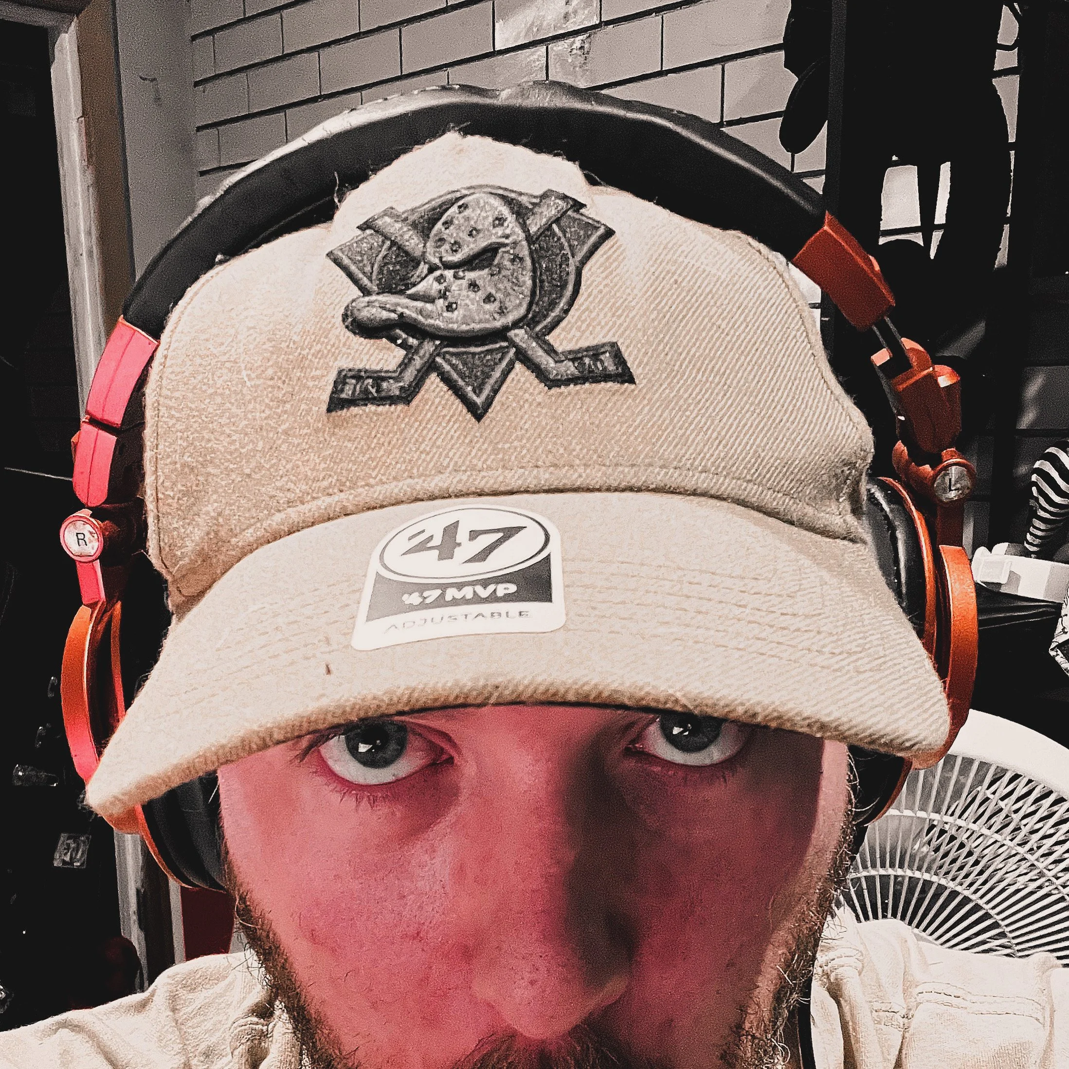 Close-up of a person wearing a baseball cap with a hockey team logo and a sticker that says '47 MVP Adjustable', and large orange and black headphones, with a fan and brick wall in the background.
