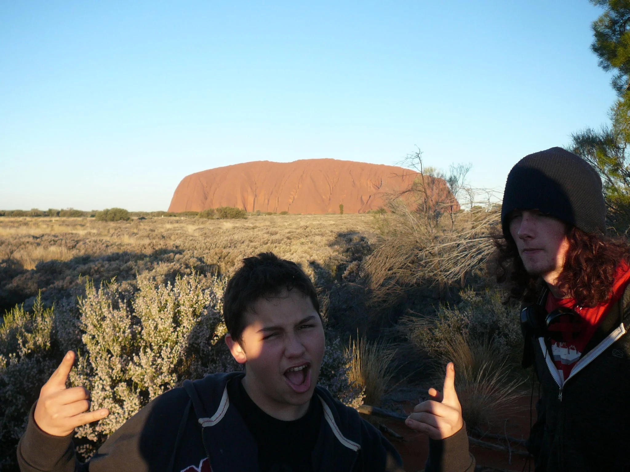 Two young men in a desert landscape with Uluru in the background during sunset. One is making a rock on hand gesture, and the other is pointing. The scene includes dry bushes and clear sky.
