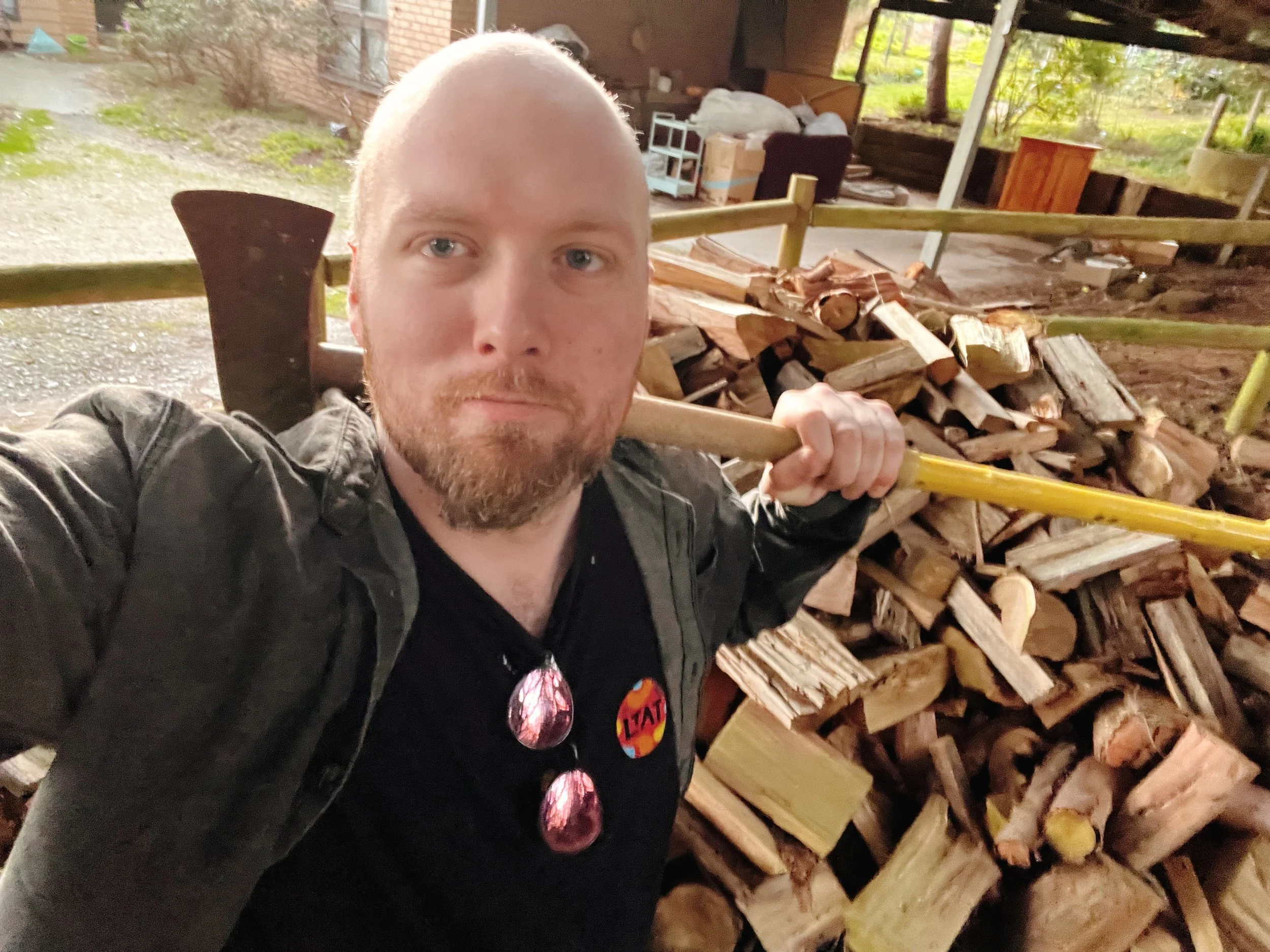 A man with a beard and sunglasses hanging from his shirt collar holding a yellow axe over his shoulder. Behind him is a large pile of chopped firewood in an outdoor shed or garage.