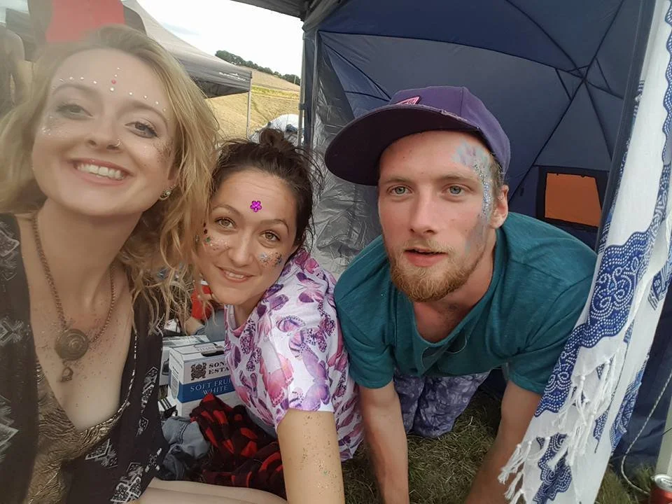 Three young adults, two women and one man, smiling and posing for a selfie at an outdoor event with tents. They have face and body glitter, and are dressed casually. One woman has a flower sticker on her forehead, and the man has face paint and is we