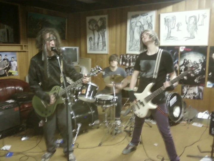 Three young men practicing rock band in a room with wooden walls, posters, and artwork.