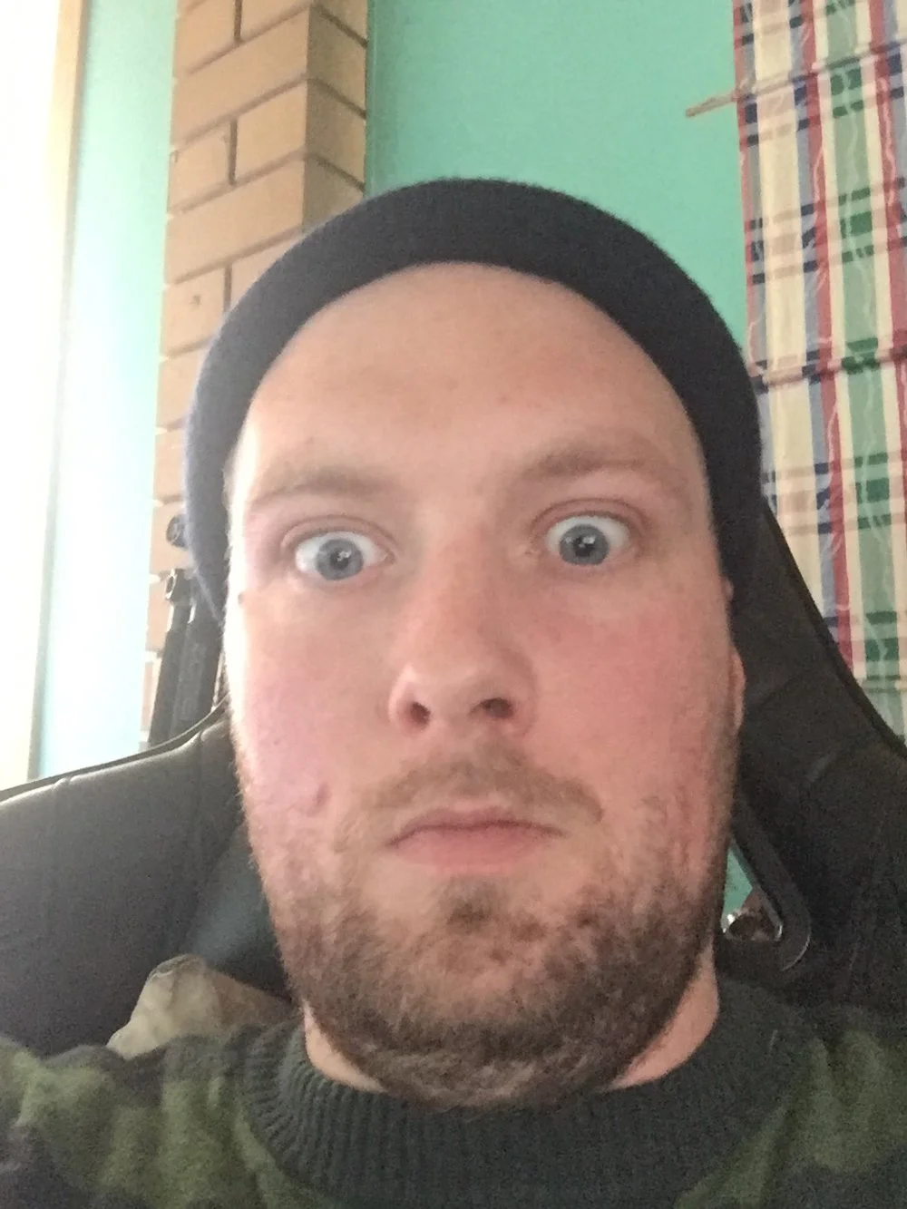 A young man with a beard and blue eyes wearing a black beanie, sitting in a room with a brick wall, green wall, and a colorful checkered curtain in the background, looking surprised.