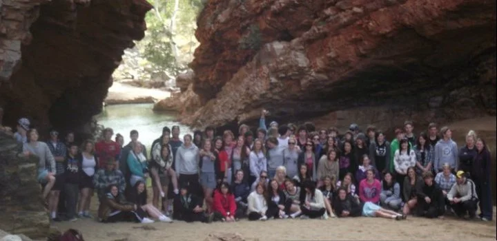Large group of people posing for a photo inside a narrow canyon with high red rock walls and a shallow water stream running through the area.