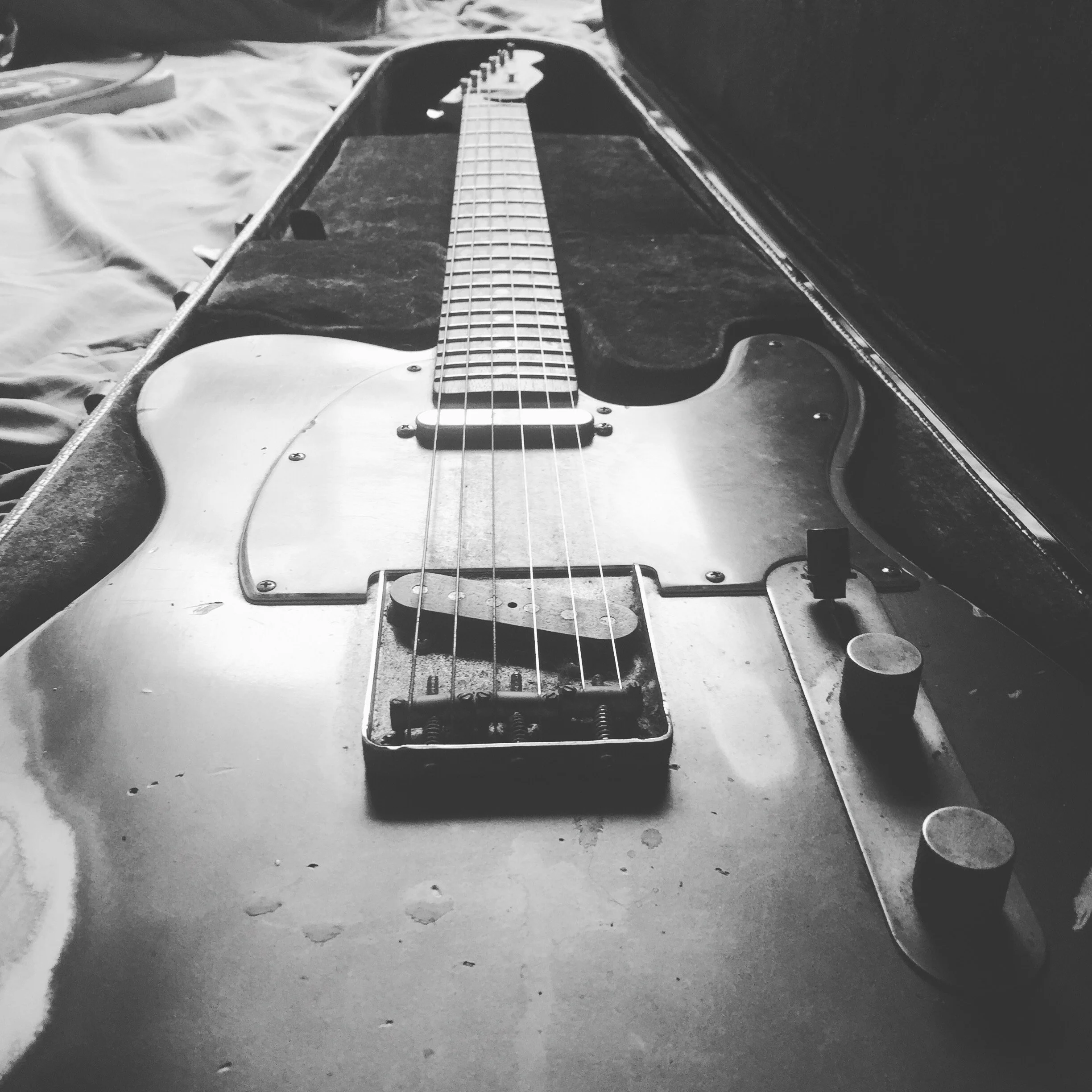 A black and white photograph of an electric guitar inside its open, plush-lined case. The guitar has a worn, solid body with six strings, a single pickup, and control knobs. The case is placed on a surface with some objects nearby.