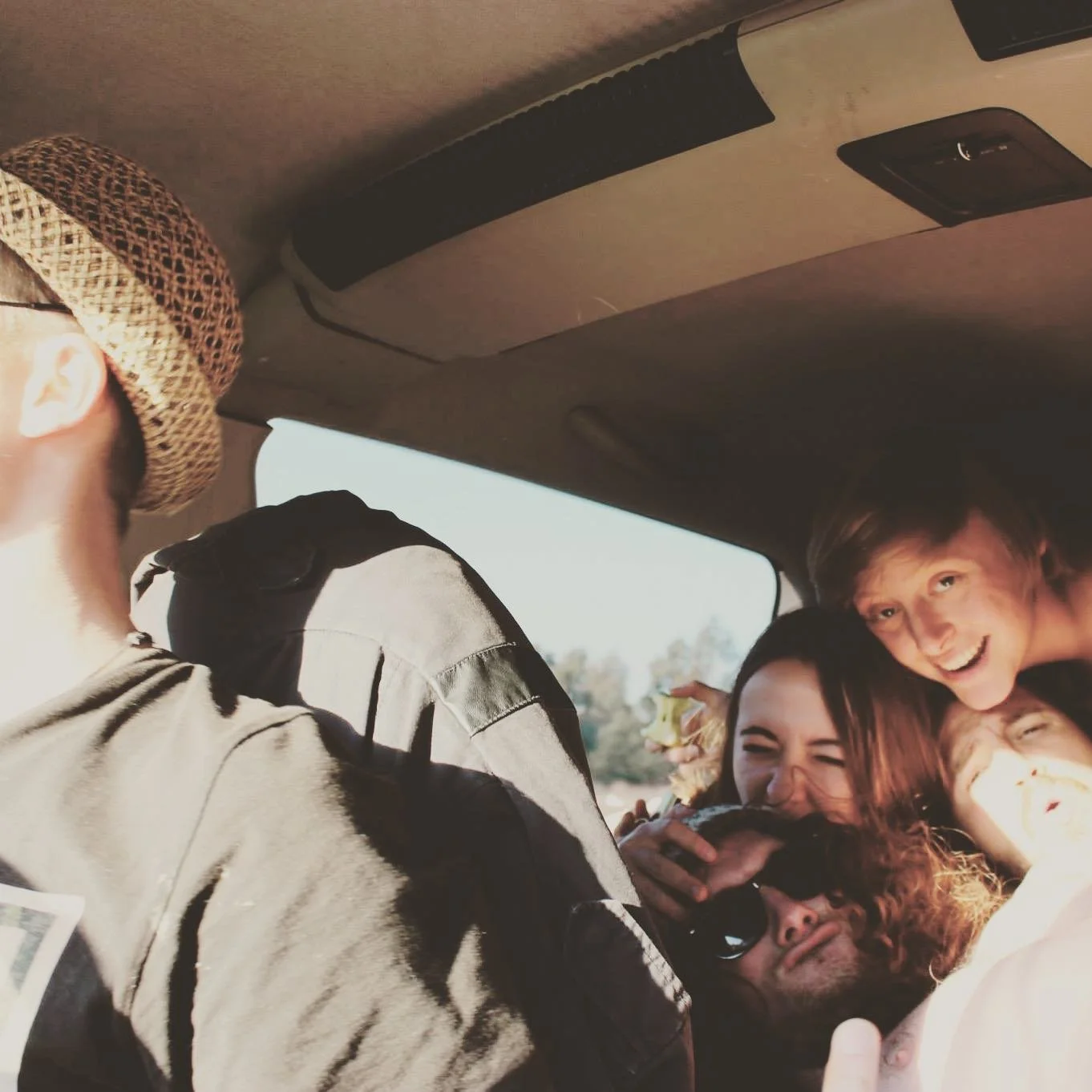 Group of young friends smiling and huddling together inside a vehicle, with some wearing sunglasses and one wearing a straw hat.