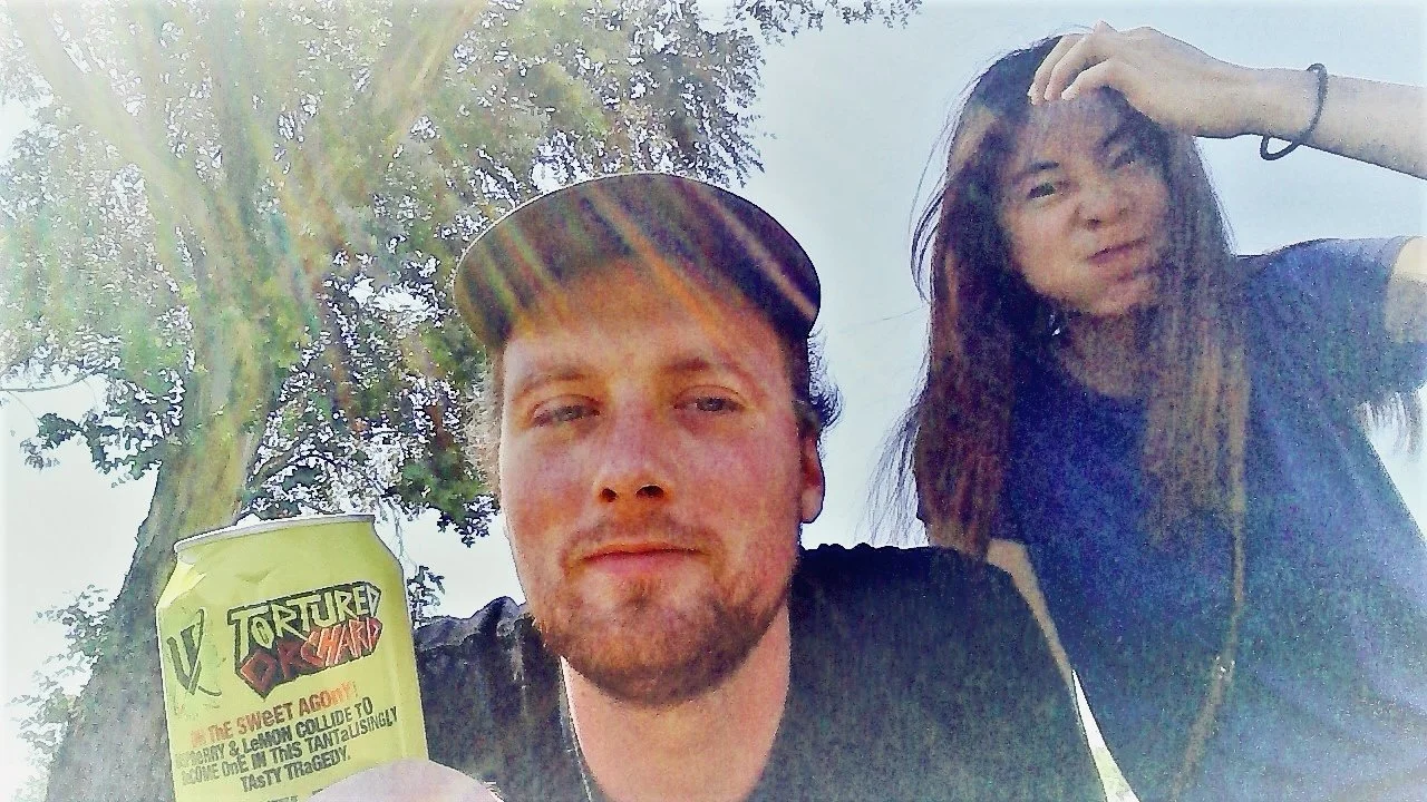 Two young adults outdoors; a man with light skin, a beard, wearing a black cap and black shirt, holding a green can of Tortured Dad soda; a woman with fair skin and long brown hair, wearing a black shirt, touching her hair, both smiling faintly. Tree