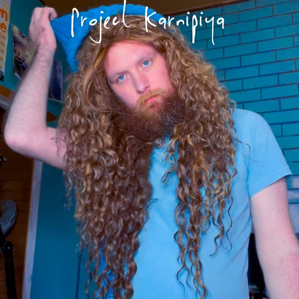 A man with long, curly red hair and a beard wearing a light blue shirt, holding a blue towel over his head in front of a blue brick wall, with the text 'Project Karnipya' written at the top.