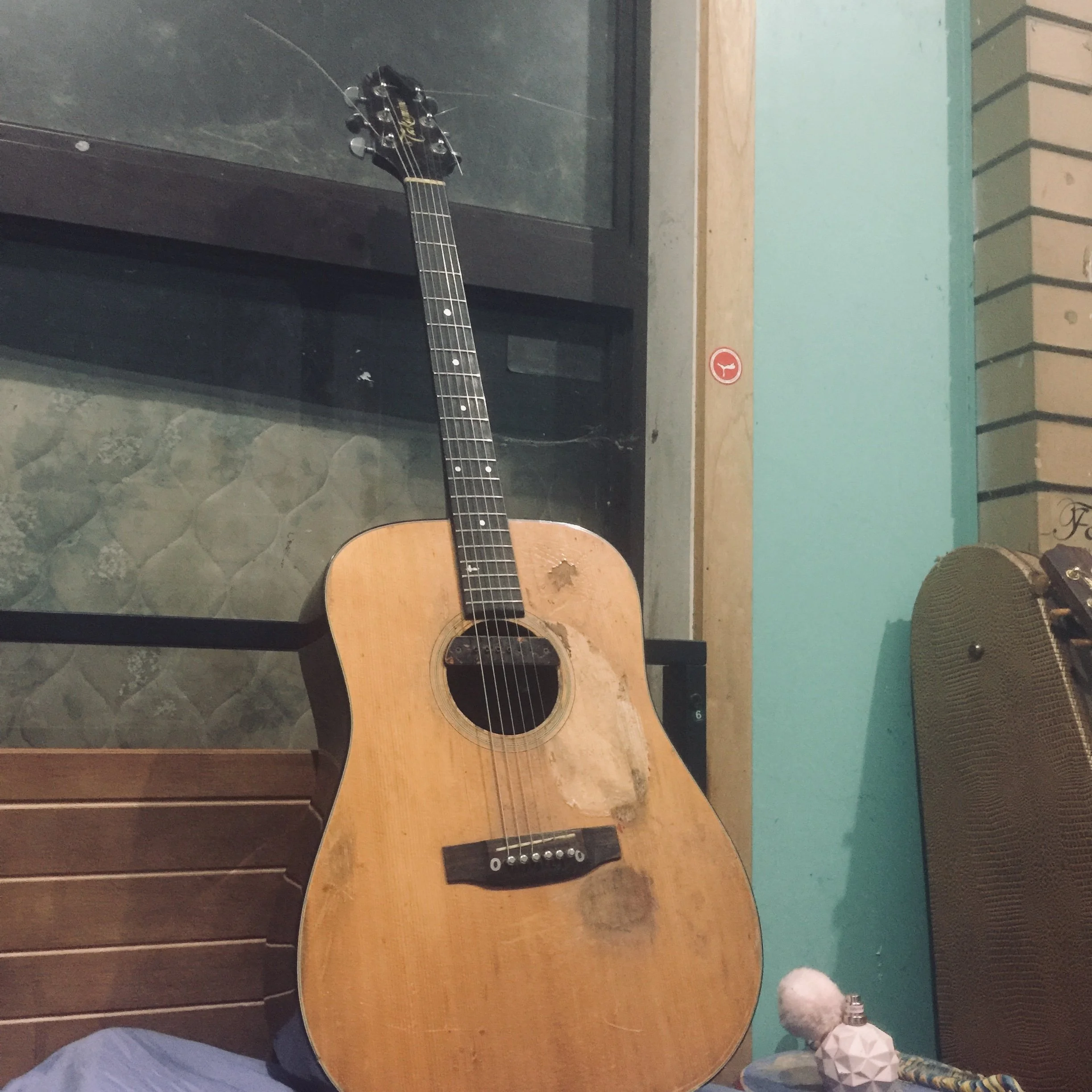An old, worn acoustic guitar with visible scratches and damage on the body, leaning against a dark wall near a window, with a small round white decorative object and a patterned cloth on a nearby surface.