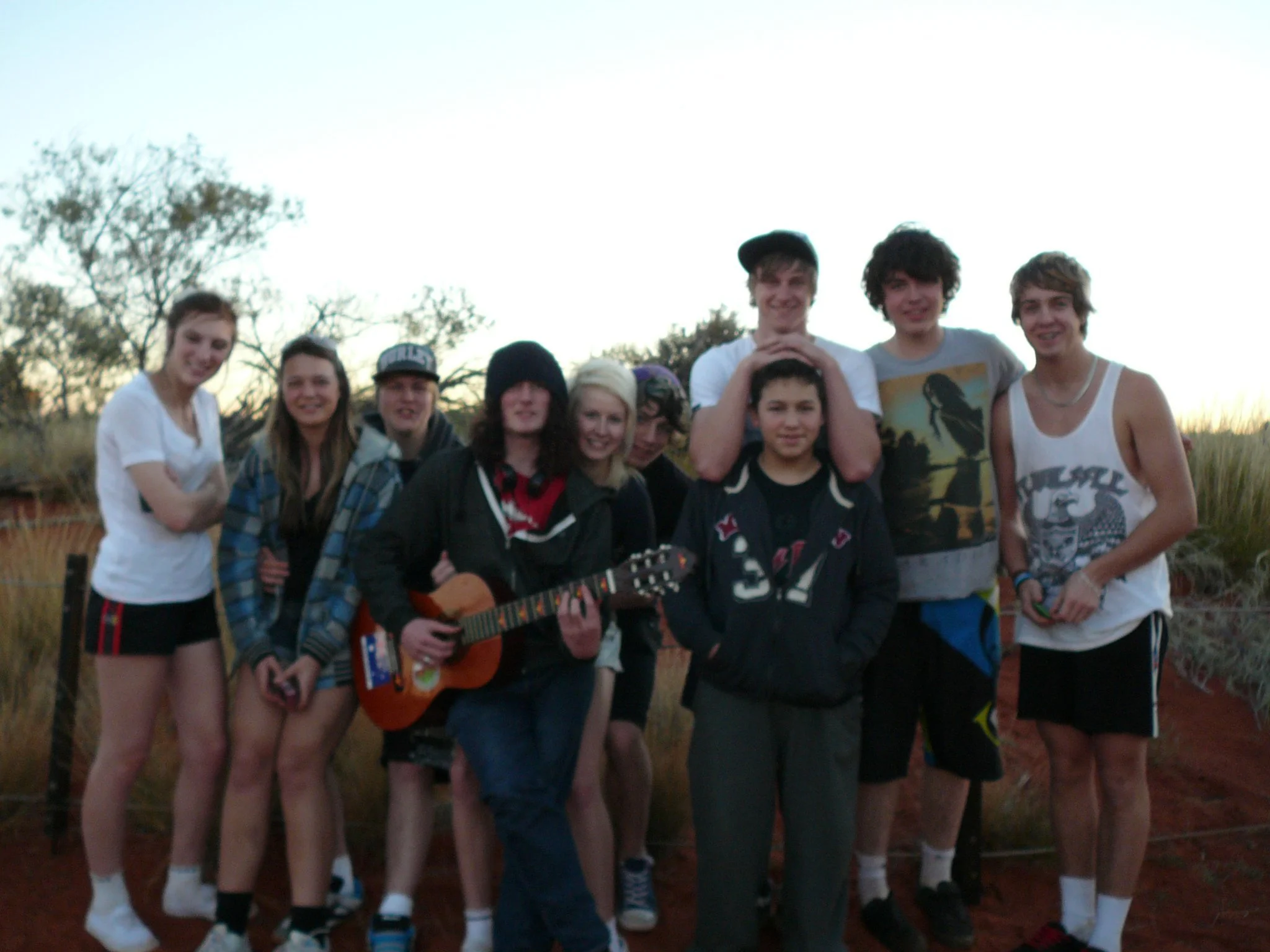 Group of young people outdoors with one person playing guitar, sunset in background, casual clothing, smiling.