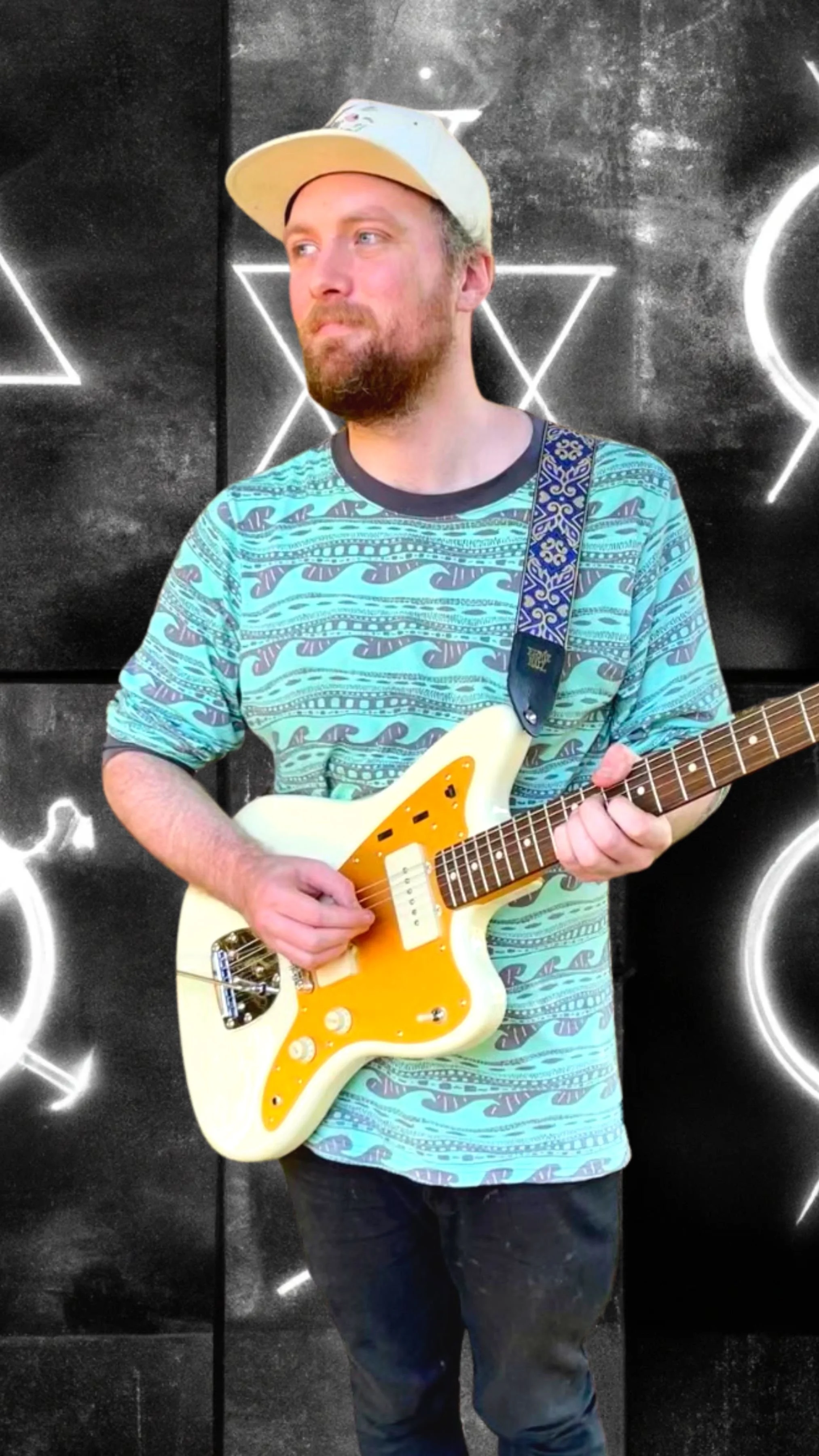 A man with a beard wearing a white cap and a turquoise patterned shirt, holding an electric guitar, standing in front of a black wall with white neon geometric shapes.